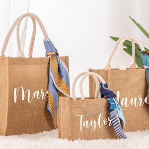 May include: Three brown burlap tote bags with white personalized names. The bags have white cotton handles and are decorated with colorful scarves.