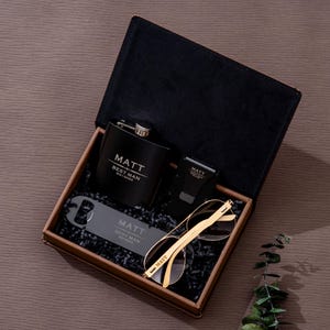 May include: A black gift box containing a flask, sunglasses, bottle opener, and a watch. The flask, bottle opener, and watch are engraved with the name "MATT" and "BEST MAN". The sunglasses have gold frames.