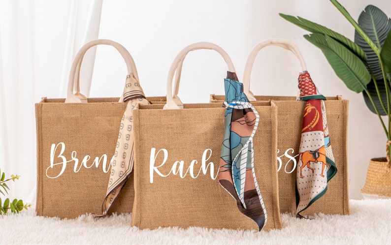 May include: Three brown burlap tote bags with white handles and personalized names: Brenda, Rachel, and Jess. Each bag has a colorful patterned scarf tied around the handle.