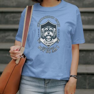 May include: A light blue t-shirt featuring a dog illustration within a shield, with the text "Neighborhood Watch" above and "Fur Real But So Am I" below. The shirt is made of a soft material.