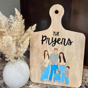 Op de afbeelding: Houten snijplank met een handvat, met een familieportret en de naam "The Pryers" in zwart schrift. De familie-illustratie bevat figuren in spijkerbroeken en diverse tops. Links staat een witte vaas met gedroogde bloemen.