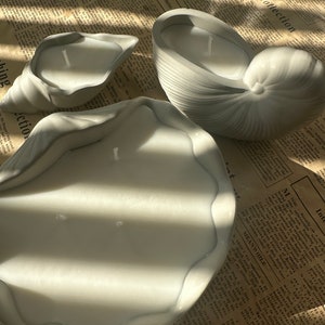 May include: Three gray seashell-shaped candles, two are smaller and one is larger. The candles are on a brown and white patterned surface.