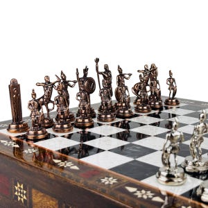 May include: A chess set with bronze-colored pieces depicting ancient warriors. The board is black and white with a decorative wooden frame. The pieces are detailed and arranged on the board, ready for play.