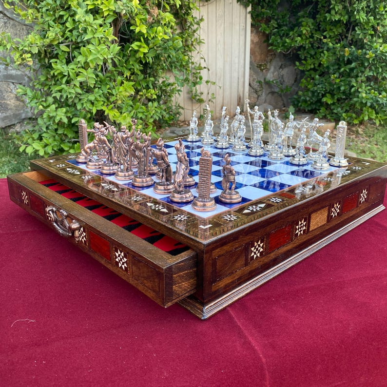 May include: A detailed chess set with a wooden board and drawer. The board features a blue and white checkered pattern with decorative trim. The chess pieces are bronze and clear glass. The drawer is open, revealing a red felt interior.