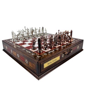 May include: A detailed chess set with silver and bronze-colored pieces on a wooden board. The board has a red and white checkered pattern and is housed in a decorative wooden box. The box has a drawer with a gold nameplate that reads "Bobby Fischer".