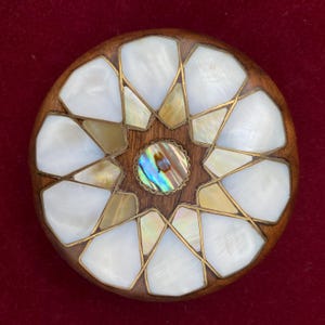 Op de afbeelding: Een ronde houten doos met een stervormig ontwerp ingelegd met parelmoer en messing. Het midden van de ster heeft een kleine, iriserende abalone-schelp.