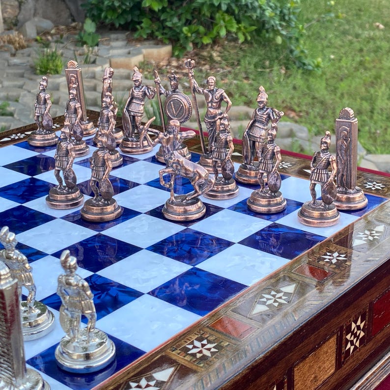 May include: A detailed chess set with bronze-colored pieces depicting ancient warriors. The chessboard features a blue and white checkered pattern, set within a decorative wooden frame. The pieces are arranged on the board, ready for play.