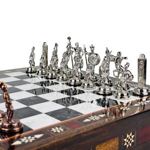 May include: A detailed chess set with silver-colored pieces depicting ancient warriors. The chessboard has a black and white checkered pattern, set on a wooden base with decorative inlays. The pieces are intricately designed.