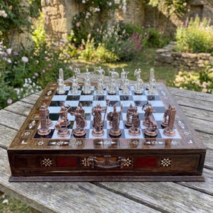 May include: A chess set with a wooden board and bronze and silver-colored pieces. The board has a black and white checkered pattern and is inlaid with decorative elements. The chess pieces are detailed, depicting figures from Greek mythology.