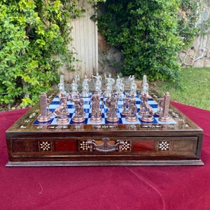 May include: A detailed chess set with bronze and silver-colored pieces arranged on a blue and white checkered board. The wooden board has a drawer and is decorated with inlaid star patterns. The chess pieces depict classical figures.