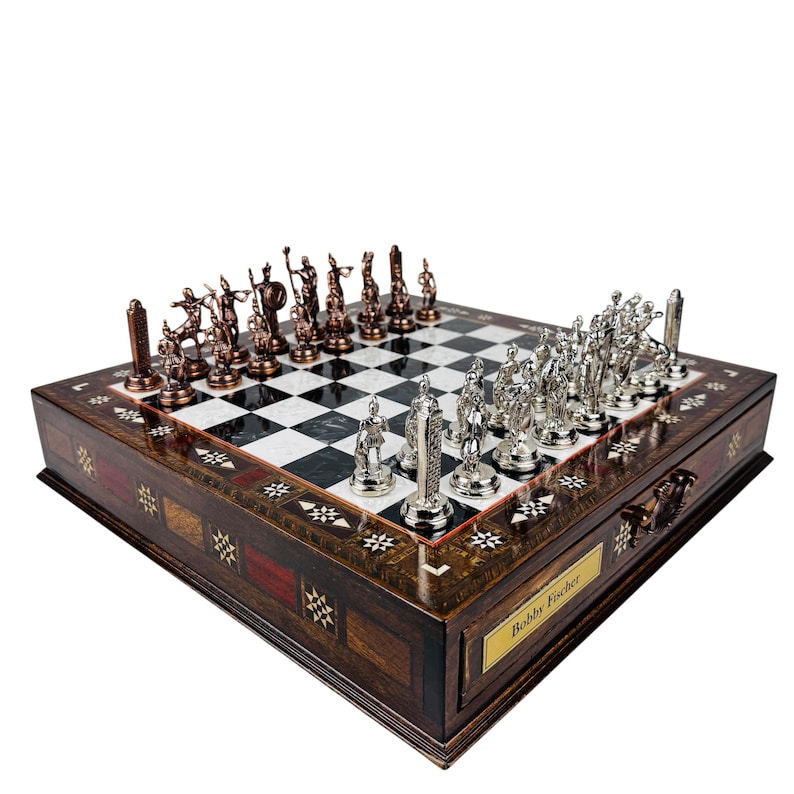 May include: A detailed chess set with bronze and silver pieces on a checkered board. The wooden box is inlaid with decorative patterns and features a drawer with a nameplate that reads "Bobby Fischer".