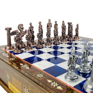 May include: A detailed chess set with bronze-colored pieces depicting classical figures. The chessboard has a blue and white checkered pattern with a decorative wooden border. The set is displayed on a white background.