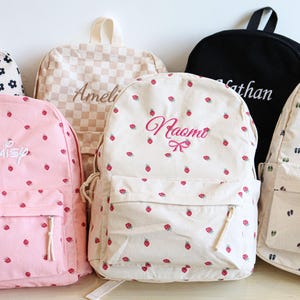 Puede incluir: Una colección de mochilas personalizadas en varios diseños. Una mochila rosa presenta un estampado de fresas y el nombre "Naomi". Otras mochilas incluyen estampados florales, a cuadros y de huellas, con nombres como "Daisy", "Ameli", "Nathan" y "James".
