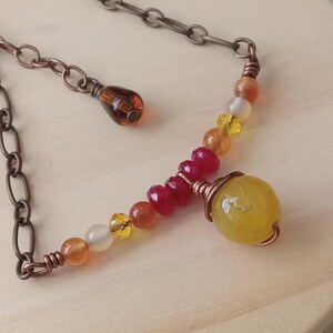 May include: A handmade necklace with a variety of colourful beads in yellow, orange, red, and white. The necklace has a copper-coloured chain and wire details, with a large yellow bead as a focal point. The necklace is displayed on a light wooden surface.