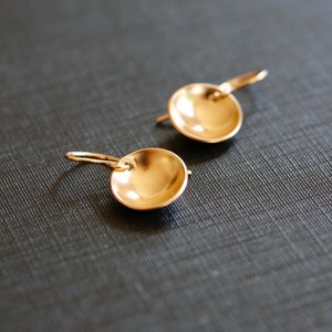 May include: A pair of gold-toned earrings with a round, slightly concave disc shape. The earrings have a simple, minimalist design.