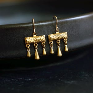 May include: A pair of gold-toned drop earrings featuring a rectangular bar with an embossed floral pattern. Each earring has three small, smooth, teardrop-shaped charms hanging from the bottom of the bar, suspended by thin metal hooks.