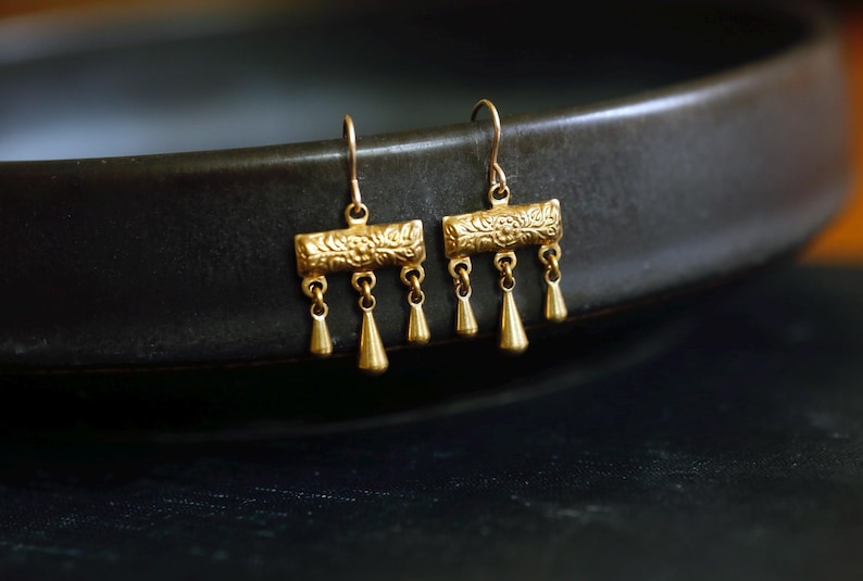 May include: A pair of gold-toned drop earrings featuring a rectangular bar with an embossed floral pattern. Each earring has three small, smooth, teardrop-shaped charms hanging from the bottom of the bar, suspended by thin metal hooks.