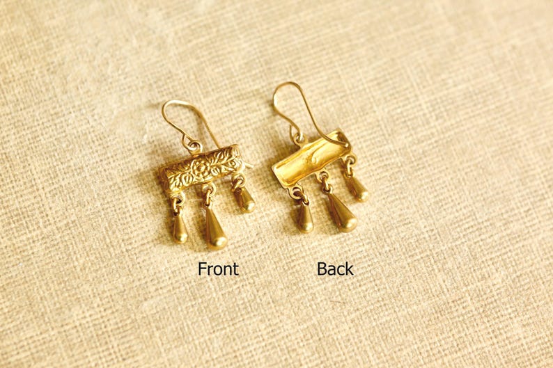 May include: A pair of gold-toned metal earrings featuring a rectangular bar with an embossed floral pattern and three dangling teardrop-shaped charms. One earring shows the detailed front, while the other displays the smooth, plain back side.