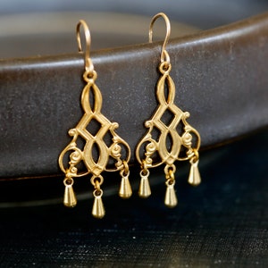 May include: Gold dangle earrings with a delicate, intricate design. The earrings feature a geometric pattern with a drop-shaped pendant.