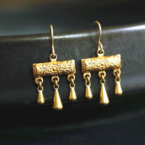 May include: A pair of gold-toned drop earrings featuring a rectangular bar with an embossed floral pattern. Each earring has three small, smooth, teardrop-shaped charms suspended from the bottom edge, attached by thin metal jump rings.