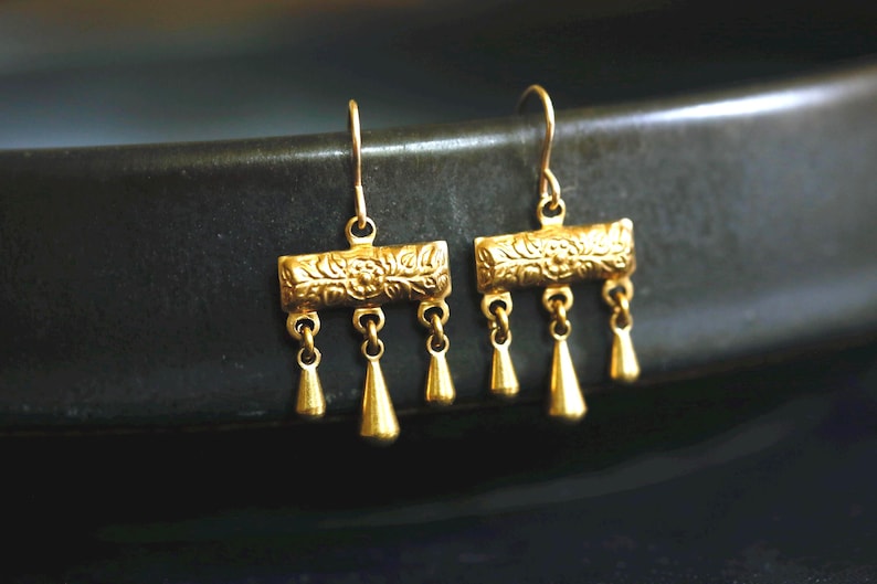 May include: A pair of gold-toned drop earrings featuring a rectangular bar with an embossed floral pattern. Each earring has three small, smooth, teardrop-shaped charms suspended from the bottom edge, attached by thin metal jump rings.