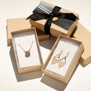 Op de afbeelding: Drie kleine kartonnen doosjes met witte binnenkant. Het bovenste doosje is vastgebonden met een zwart lint en heeft een zwart-wit patroon. De andere twee doosjes bevatten sieraden: een gouden ketting met een groene edelsteen hanger en gouden oorbellen met een geometrisch ontwerp.