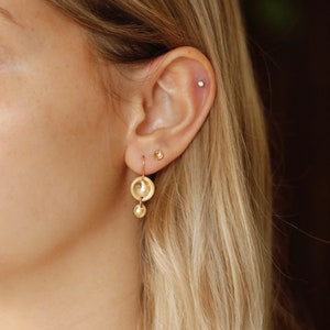 May include: A pair of gold earrings with a small round disc and a larger round disc hanging from a thin gold chain. The earrings are worn in the left ear of a person with blonde hair.