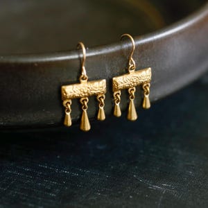 May include: A pair of gold-toned drop earrings featuring a rectangular bar with an embossed floral pattern. Each earring has three small, dangling teardrop-shaped charms suspended from the bottom edge, attached to a simple curved ear wire hook.
