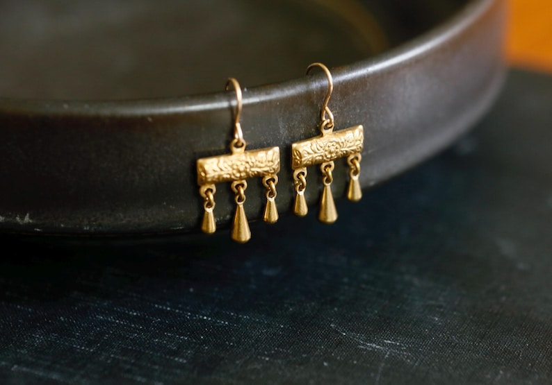 May include: A pair of gold-toned drop earrings featuring a rectangular bar with an embossed floral pattern. Each earring has three small, dangling teardrop-shaped charms suspended from the bottom edge, attached to a simple curved ear wire hook.