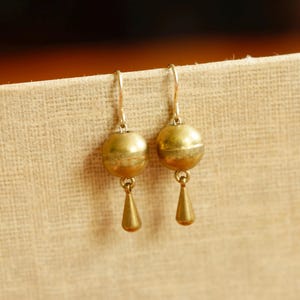May include: A pair of gold-colored earrings. Each earring features a round, ball-shaped top with a small, teardrop-shaped pendant hanging below. The earrings are suspended from a simple hook.