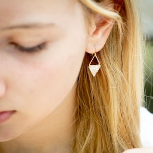May include: A pair of gold triangle earrings with white beads. The earrings are hanging from the earlobe of a person with blonde hair.