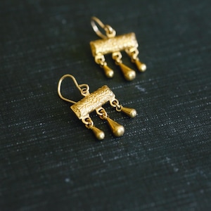May include: A pair of gold-toned drop earrings featuring a rectangular bar with an intricate embossed pattern. Each bar is suspended from a curved ear wire and features three small, dangling teardrop-shaped charms hanging from the bottom edge.