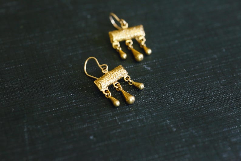 May include: A pair of gold-toned drop earrings featuring a rectangular bar with an intricate embossed pattern. Each bar is suspended from a curved ear wire and features three small, dangling teardrop-shaped charms hanging from the bottom edge.