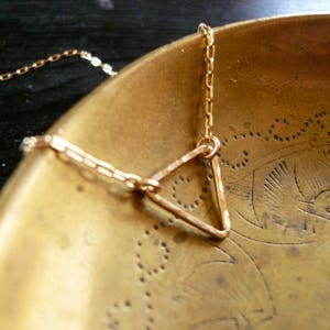 May include: A gold triangle pendant necklace with a delicate chain. The necklace is displayed on a brass bowl with an intricate design.
