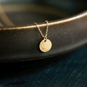 May include: A gold necklace with a hammered, round pendant. The necklace is on a dark brown surface.