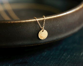 May include: A gold necklace with a hammered, round pendant. The necklace is on a dark brown surface.