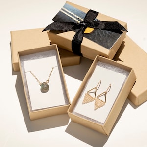 May include: A gold chain necklace with a round pendant and a pair of gold triangle-shaped drop earrings with beaded detail. The jewelry is displayed on white cards inside two open brown cardboard boxes, with a third closed box tied with a black ribbon.