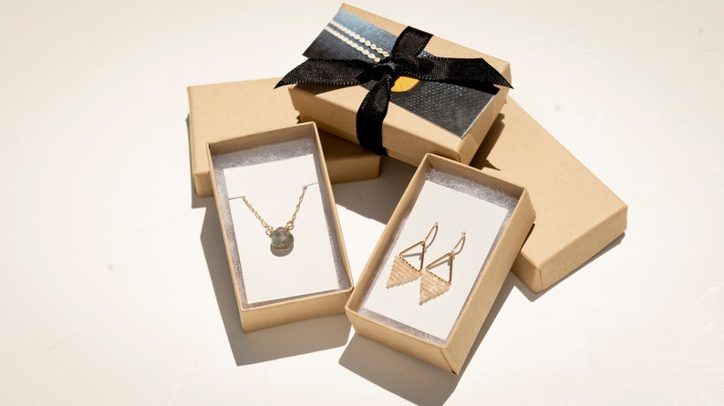 May include: A gold chain necklace with a round pendant and a pair of gold triangle-shaped drop earrings with beaded detail. The jewelry is displayed on white cards inside two open brown cardboard boxes, with a third closed box tied with a black ribbon.