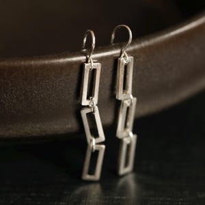 May include: Silver rectangular dangle earrings. Each earring features three connected open rectangles. The earrings are suspended from a simple hook. The jewelry is displayed against a dark background.