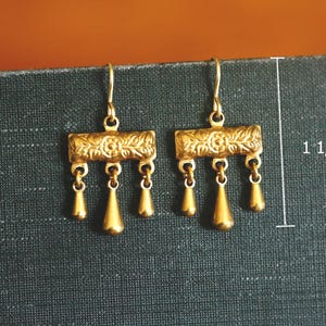 May include: A pair of gold-toned drop earrings featuring a rectangular bar with an embossed floral pattern and three dangling teardrop charms. The earrings measure 1 1/8 inches in total length and are suspended from simple curved ear wires.