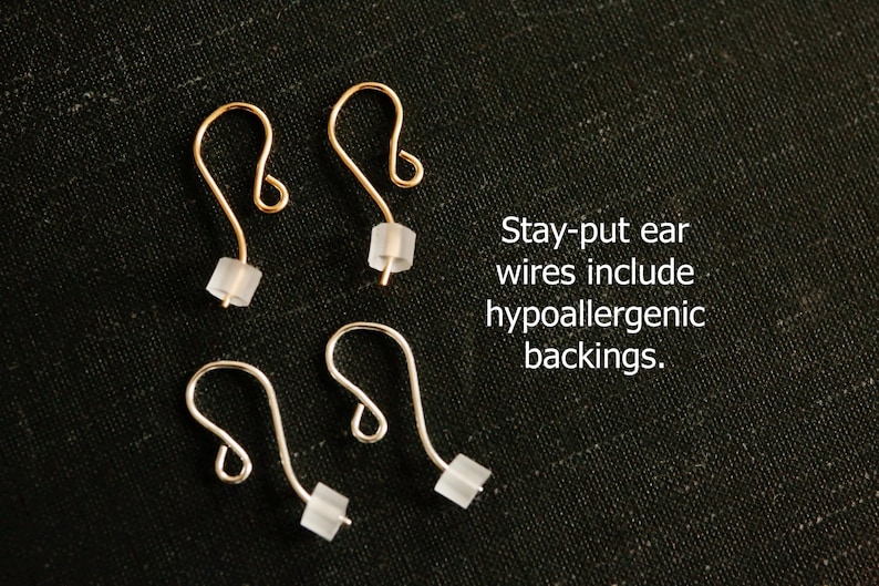 May include: Two pairs of ear wires, one gold-toned and one silver-toned, each featuring a curved hook design and a small, translucent, hypoallergenic backing. The text reads: Stay-put ear wires include hypoallergenic backings.