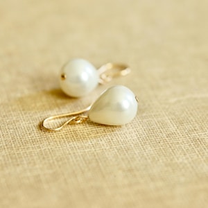 May include: A pair of elegant pearl earrings with gold-colored hooks. One earring features a round pearl, while the other has a teardrop-shaped pearl. The earrings are set against a textured, light brown background.