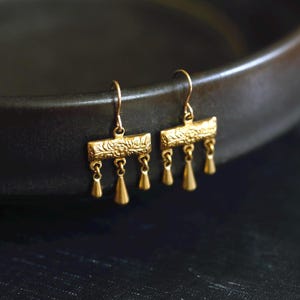 May include: A pair of gold-toned drop earrings featuring a rectangular bar with an embossed floral pattern. Three small, smooth, teardrop-shaped charms hang from the bottom of each bar, suspended by thin metal loops attached to curved ear wires.