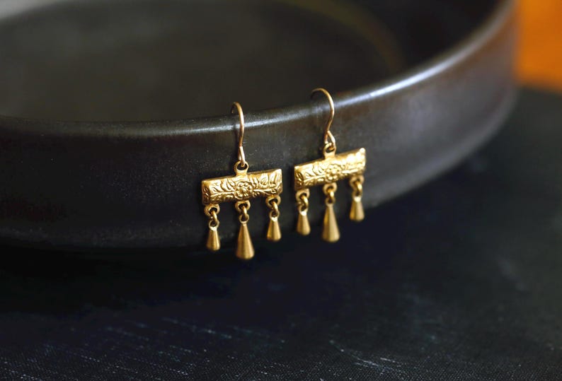 May include: A pair of gold-toned drop earrings featuring a rectangular bar with an embossed floral pattern. Three small, smooth, teardrop-shaped charms hang from the bottom of each bar, suspended by thin metal loops attached to curved ear wires.