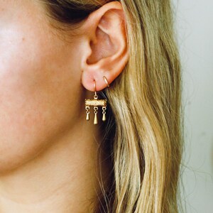 May include: A gold-toned drop earring featuring a horizontal cylindrical bar with an intricate embossed pattern. Three small, teardrop-shaped charms hang from the bottom of the cylinder by delicate links, suspended from a simple gold-toned ear wire hook.