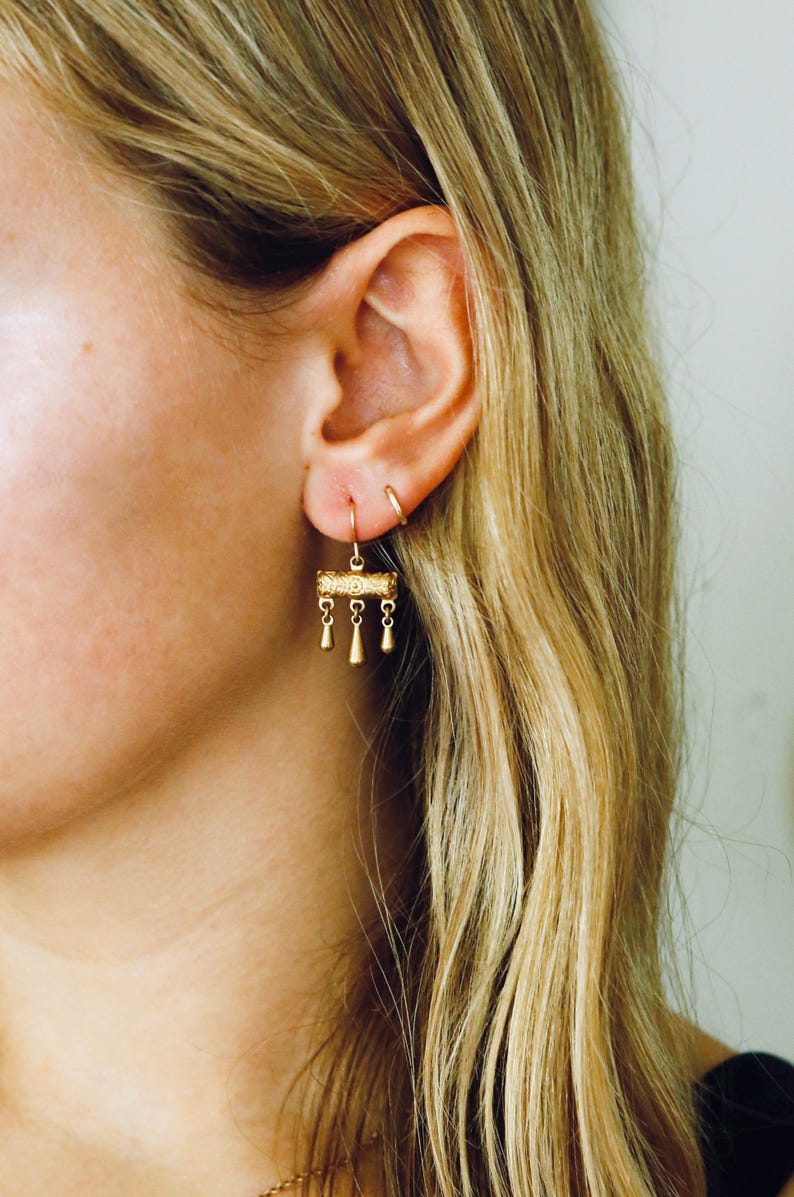 May include: A gold-toned drop earring featuring a horizontal cylindrical bar with an intricate embossed pattern. Three small, teardrop-shaped charms hang from the bottom of the cylinder by delicate links, suspended from a simple gold-toned ear wire hook.