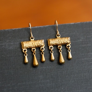 May include: A pair of gold-toned drop earrings featuring a rectangular bar with an embossed floral pattern. Each earring has three small, smooth, teardrop-shaped charms suspended from the bottom edge, attached to a curved ear wire hook.