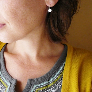 May include: A woman wearing a gray shirt with embroidered details and a yellow cardigan. She is wearing small silver earrings.