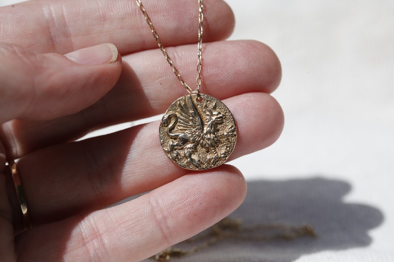 Op de afbeelding: Een gouden ketting met een ronde hanger met een gedetailleerd, reli&euml;fbeeld van een griffin.