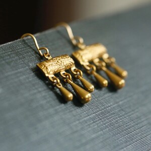 May include: A pair of gold-toned drop earrings featuring a rectangular top bar with an embossed floral pattern. Three small, smooth, teardrop-shaped charms hang from each bar, suspended by thin metal loops. The earrings include curved wire hooks.